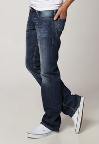 Jeans de mezclilla de color azul oscuro con un corte relajado, detalles de desvanecimiento y costuras en contraste. Combinados con zapatillas blancas para un look casual.
