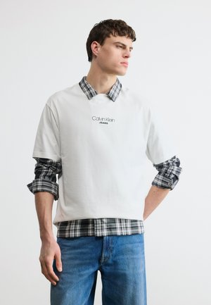 Mladý muž nosiaci biele tričko Calvin Klein Jeans cez čierno-bielu károvanú košeľu a modré džínsy, pozerajúci sa do strany.