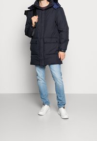 Parka imbottito blu navy con cappuccio, dotato di zip frontale, bottoni a pressione, due tasche frontali e polsini elasticizzati; indossato sopra jeans azzurri e sneakers bianche.