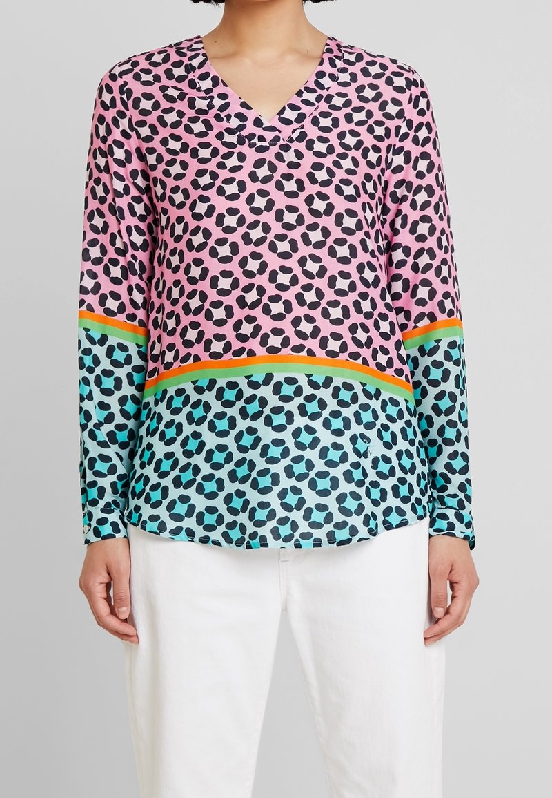 Femme portant un haut à manches longues avec col en V imprimé léopard rose et bleu séparé par des rayures orange et vertes, associé à un pantalon blanc.