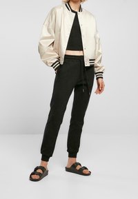 Veste bomber en satin beige clair avec des rayures noires, associée à un pantalon de survêtement noir et des sandales foncées. Coupe décontractée avec un design raccourci.