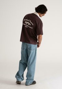 Mand med krøllet hår iført en mørk oversized t-shirt med hvid tekst og lyseblå baggy jeans, stående mod en simpel lys baggrund.
