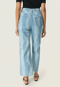 Jeans de tiro alto de color azul claro con un patrón ondulado, corte de pierna ancha y dos bolsillos traseros. Llevados con una blusa negra y sandalias con tiras.