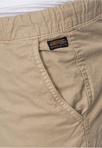 Pantalon en coton beige avec une taille élastique, poche avant visible et une petite étiquette bleu foncé avec du texte orange au-dessus de la poche.