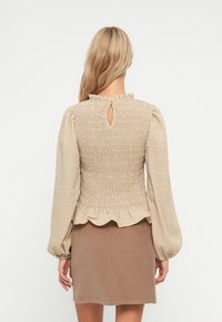 Beige smocked tops ar ar izplūdušo apakšmalu, atslēgas cauruma aizmugures detaļu un garām uzpūstām piedurknēm. Nēsāts virs brūnas pieguļošas svārku. Tekstūrēta auduma.