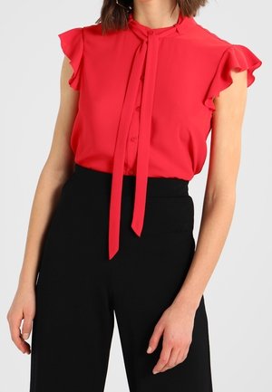 Vrouw draagt een rood mouwloos bloesje met gerimpelde schouders en een strik aan de voorkant, gecombineerd met hooggesneden zwarte broek.