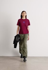 Βουργουνδί φανελάκι με μπλε λογότυπο, συνδυασμένο με χαλαρούς ελαιόχρωμους παντελόνες και μαύρες μπότες μέχρι τον αστράγαλο. Το μοντέλο κρατάει μια μαύρη δερμάτινη ζακέτα.