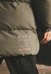 Giacca imbottita in tessuto verde oliva con coordinate stampate, dettagli in edizione limitata e cuciture texturizzate. Pantaloni neri parzialmente visibili.