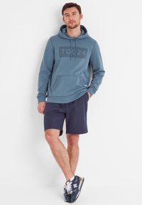 Blue hoodie with a grey "TOG24" logo, kangaroo pocket, and drawstring hood, paired with dark blue shorts and navy sneakers.