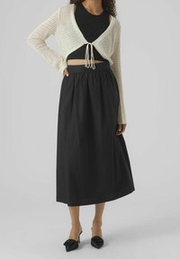 Žena v krémovém háčkovaném krátkém kardiganu přes černý crop top, černé midi sukni s vysokým pasem a černých lodičkách s mašlemi.