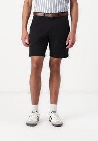 Shorts noirs en tissu léger, associés à une ceinture marron. Portés avec des sneakers blancs et gris avec des rayures noires.