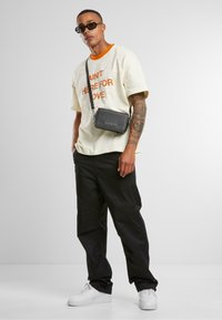 Sac banane noir, forme rectangulaire, en matériau synthétique. Le modèle porte une chemise crème avec un texte orange, un pantalon noir et des baskets blanches.