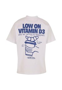 Valkoinen puuvillapaita, jossa on sininen graafinen teksti: "ALHAISET D3-VITAMIINITASOT, KORKEA KAHVI- JA CROISSANTTIEN KULUTUS" kuvitetulla kahvikupilla ja leivonnaisella.