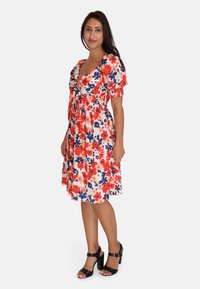 Vestido floral en crema con flores rojas y azules, mangas cortas abullonadas, cintura ajustada y falda acampanada. Sandalias de tacón alto negras.