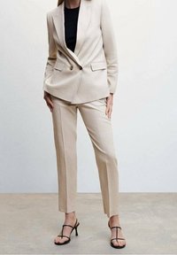 Béžový přizpůsobený oblek s dvouřadým sakem se zlatými knoflíky, v kombinaci s úzkými kalhotami; nošeno s černým topem a sandály na tenkých páscích.