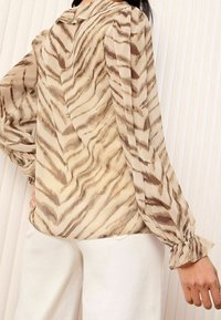 Blusa beige con strisce a tigre marroni, con colletto e polsini arruffati, tessuto morbido e maniche lunghe a sbuffo. Design etereo e fluido.