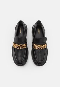 Mocassins en cuir noir avec une épaisse chaîne en or et des ornements en strass sur le dessus, marqués Guess à l'intérieur, vus de dessus sur fond blanc.