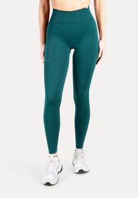 Leggings met hoge taille in zeegroen, gemaakt van rekbare stof, voorzien van een zijzak en een gladde textuur, gecombineerd met witte sportschoenen.