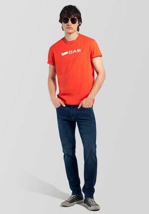 Hombre con gafas de sol negras, camiseta roja de manga corta con el logo "GAS", vaqueros azul oscuro y zapatillas negras con suelas blancas.