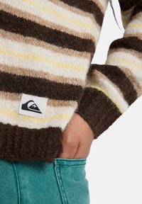 Gebreide trui met horizontale strepen in bruin, crème en geel. Kenmerkt zich door een geribde manchette en een logopatch aan de zoom. Te combineren met groene broek.