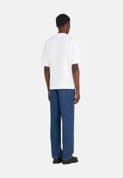 Homme debout, de dos, portant un t-shirt blanc uni à manches courtes, un jean bleu droit et des chaussures noires sur un fond blanc.