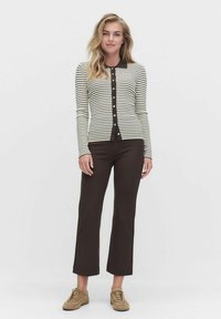 Vrouw in een crèmekleurig en zwart gestreept blouse met knopen, donkere bruine cropped broek en beige sneakers, staande tegen een witte achtergrond.