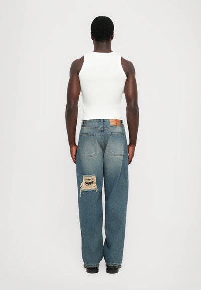 Blaue Denim-Jeans mit lockerer Passform, ausgestattet mit einem großen, distressed Patch am linken Oberschenkel und einem braunen Leder-Logoschild am hinteren Bund.