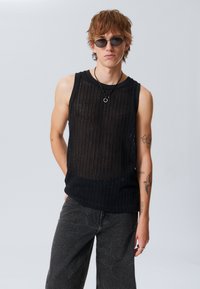 Haut sans manches en tricot noir avec des rayures verticales, associé à un pantalon texturé gris. Les accessoires comprennent des lunettes de soleil et un collier pendentif.