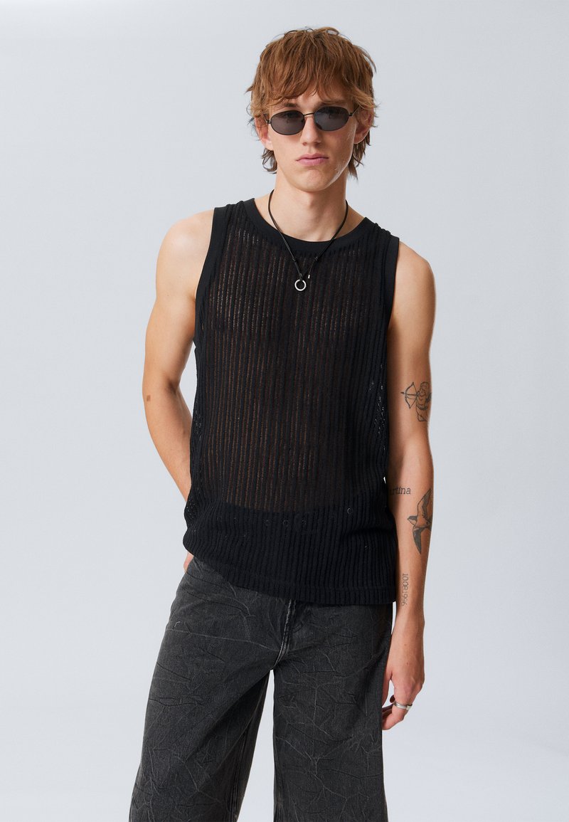 Haut sans manches en tricot noir avec des rayures verticales, associé à un pantalon texturé gris. Les accessoires comprennent des lunettes de soleil et un collier pendentif.