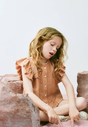Jeune fille aux cheveux ondulés portant une robe à manches à volants, assise en tailleur sur une surface rose, pointant vers le bas avec les deux mains.