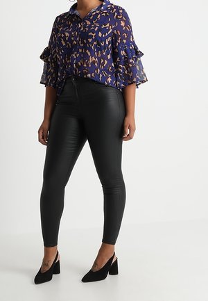 Femme portant un chemisier bleu à imprimé léopard avec des manches évasées, un pantalon en cuir noir taille haute, et des chaussures noires à talons carrés.