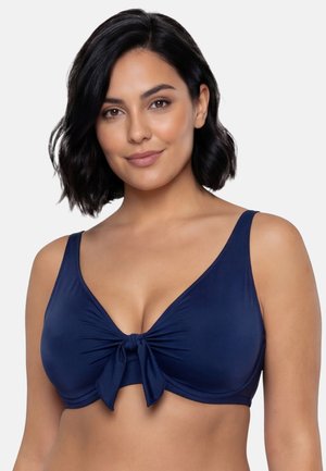 Kvinde med kort sort hår iført en mørkeblå bikinitop med knude foran, der smiler let mod en ensfarvet baggrund.
