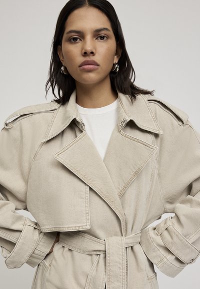 Cappotto trench in denim beige con vita regolabile, ampi revers e spalline. Presenta una silhouette rilassata e dettagli di cucitura in vista.