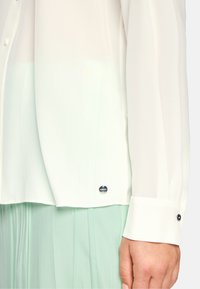 White blouse in lightweight fabric with long sleeves, featuring a buttoned cuff and a subtle logo detail near the hem.