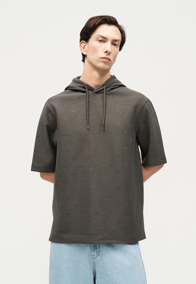 Νεαρός άνδρας στέκεται με τα χέρια πίσω από την πλάτη, φορώντας γκρι σκούρο κοντομάνικο φούτερ με διακριτικό σχέδιο και ανοιχτό μπλε τζιν παντελόνι.