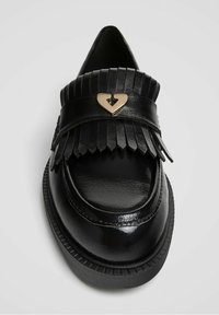 Mocassin en cuir noir avec des détails fringés, un accent doré en forme de cœur sur la bride et une semelle en caoutchouc texturé. Fini lisse et brillant.