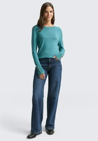 Türkisfarbener Pullover mit langen Ärmeln und Knopfakzenten an den Manschetten, kombiniert mit dunkelblauen Jeans mit weitem Bein und schwarzen Stiefeletten.