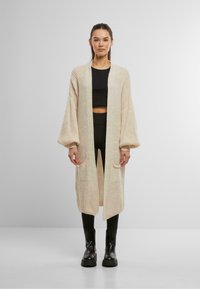 Strickjacke in Creme mit offenem Vorderteil, langen Ärmeln, Seitentaschen und lockerer Passform. Das Modell trägt dazu ein schwarzes Oberteil und eine schwarze Hose.