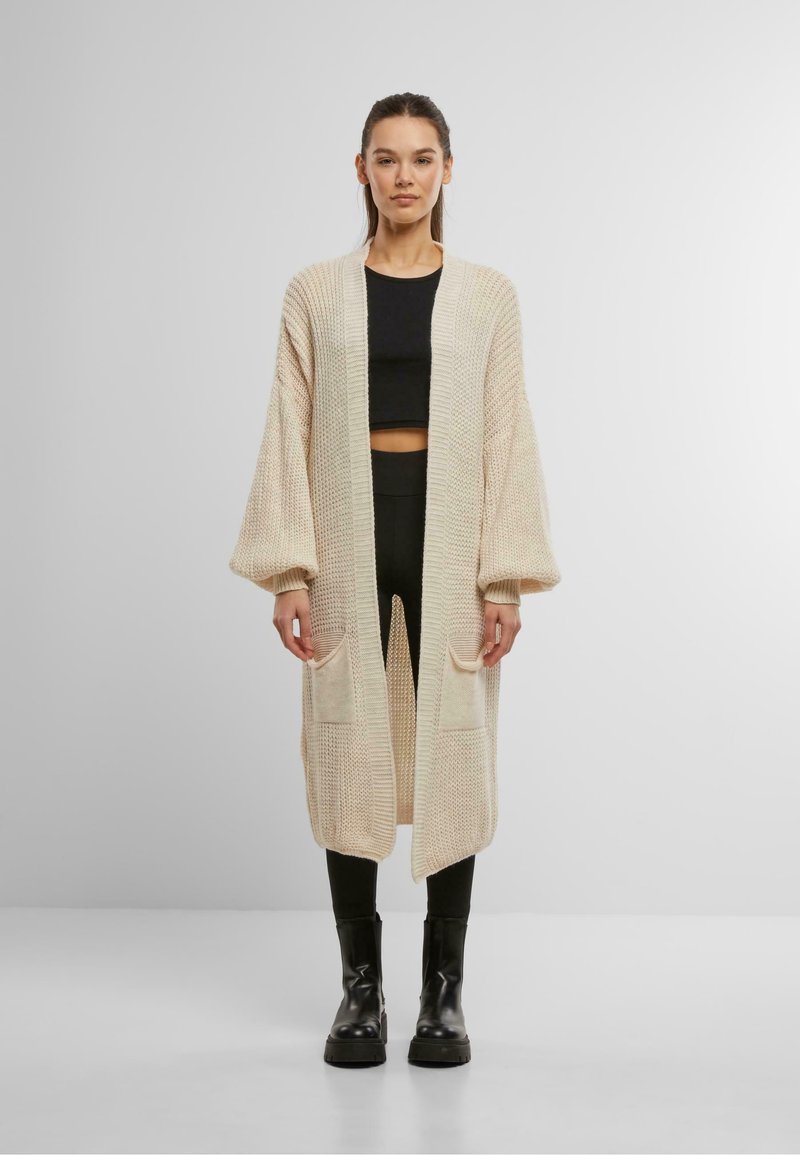 Strickjacke in Creme mit offenem Vorderteil, langen Ärmeln, Seitentaschen und lockerer Passform. Das Modell trägt dazu ein schwarzes Oberteil und eine schwarze Hose.
