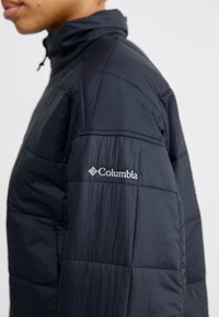Personne portant une veste matelassée noire Columbia avec le logo brodé sur le haut de la manche, vue de profil sur un fond uni.