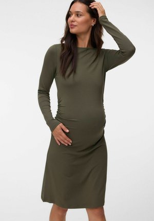 Gravid kvinde iført en olivengrøn, tætsiddende kjole med lange ærmer, med den ene hånd på maven og den anden, der rører ved hendes hår.