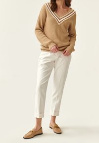 Frau in beiger V-Ausschnitt-Strickjacke mit weißen Streifen, weißen Cropped-Hosen und beigen Loafers mit goldener Schnallen-Detail.