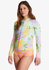Camisa de surf de manga larga con estampado tropical colorido en tonos pastel, con cremallera frontal y diseño ajustado, combinada con un pantalón de patrón de olas en amarillo y rosa.