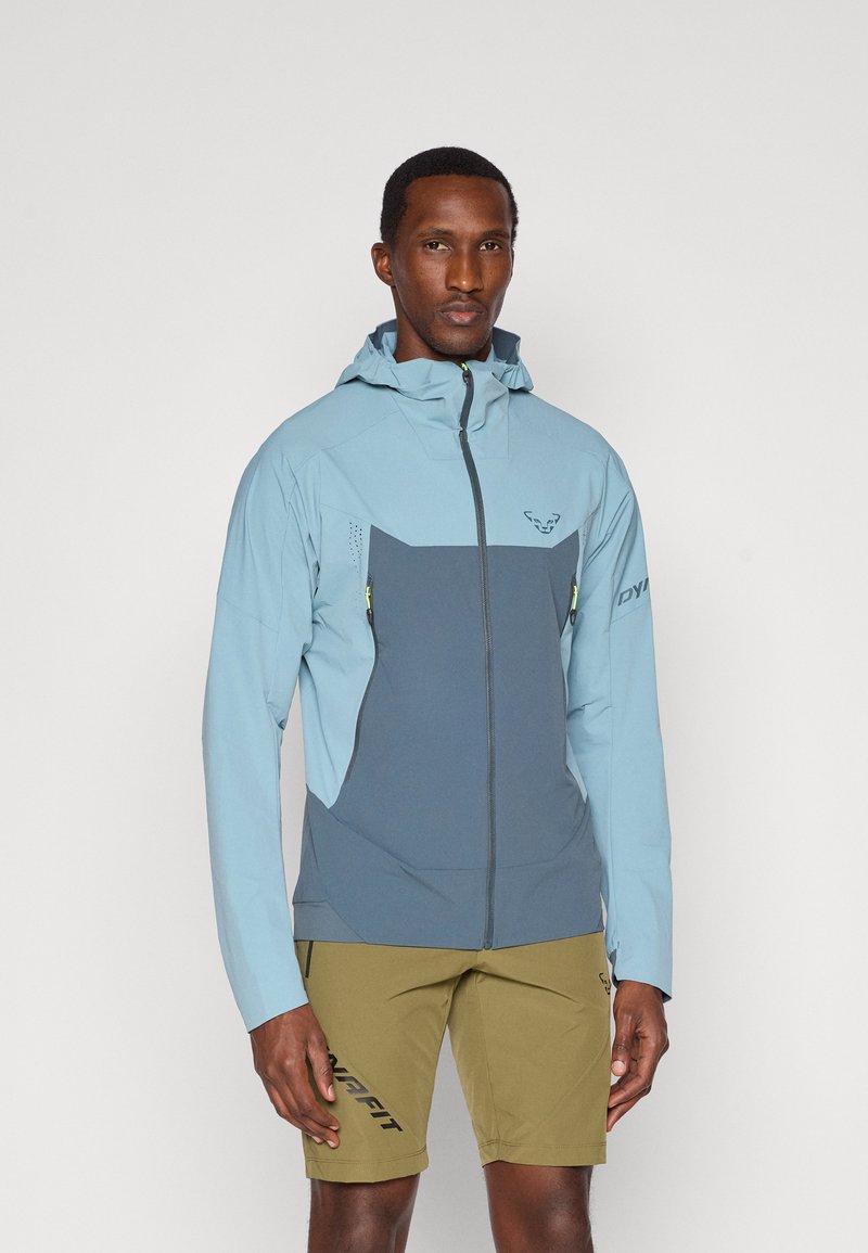 Ελαφρύ, δίχρωμο μπουφάν; το επάνω μισό είναι ανοιχτό μπλε, το κάτω μισό σκούρο γκρι; διαθέτει τσέπες με φερμουάρ και κουκούλα; κατασκευασμένο από αναπνεύσιμο υλικό.