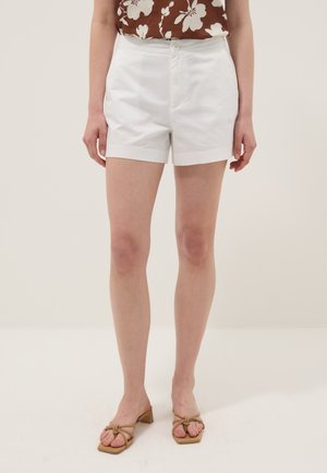 Weibliches Model trägt weiße Shorts mit hoher Taille, braunes ärmelloses Top mit Blumenmuster und beige Sandalen mit Absatz, steht vor einem schlichten hellen Hintergrund.