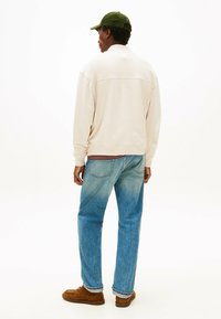 Felpa beige con polsini a coste, jeans azzurri e mocassini marroni. Vista posteriore, il modello indossa un cappello verde scuro.