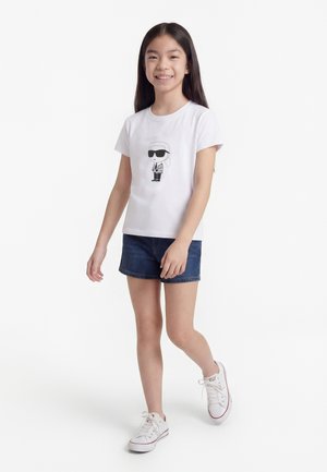 Fetiță zâmbitoare, purtând un tricou alb cu grafică, pantaloni scurți din denim închis și adidași albi, mergând cu un picior înainte pe un fundal alb simplu.