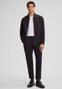 Mężczyzna stoi ubrany w czarną kurtkę zapinaną na zamek, białą koszulkę, czarne spodnie, czarne skarpetki oraz czarne sznurowane buty na jednolitym szarym tle.