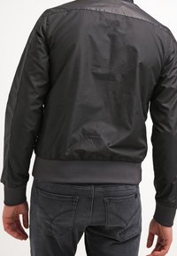 Veste bomber noire avec une texture lisse et brillante, des poignets et un ourlet côtelés, et un design épuré et simple. Présente des détails de couture subtils.