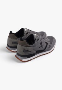Zapatillas deportivas grises confeccionadas con materiales de ante y tela, que presentan un logo de calavera negra, detalles texturizados y una suela de goma marrón claro.